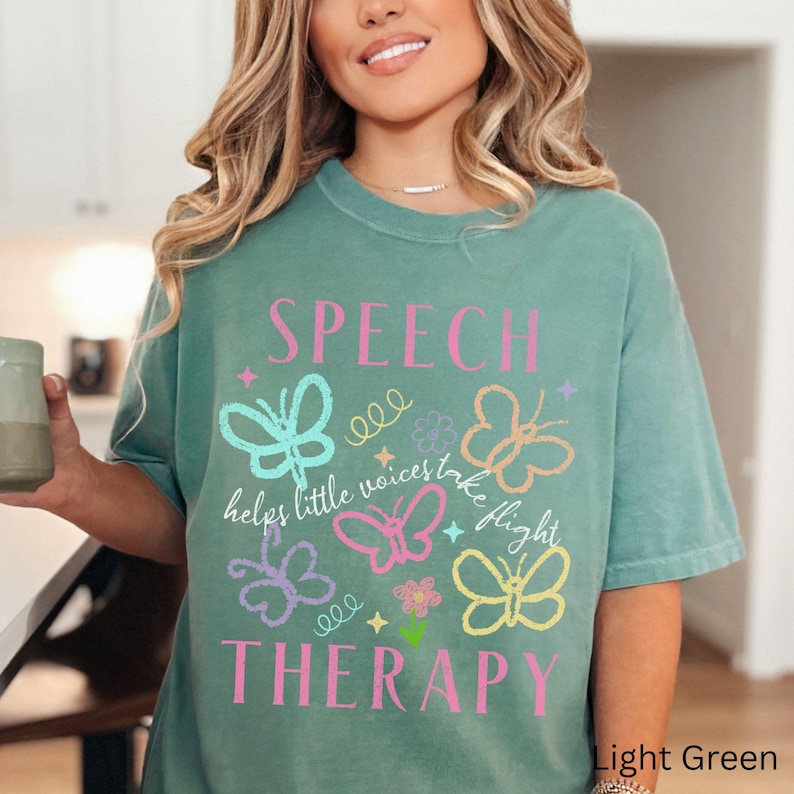 Puede incluir: Camiseta verde claro con las palabras "SPEECH THERAPY" y la frase "helps little voices take flight". El dise&ntilde;o incluye mariposas y flores coloridas.