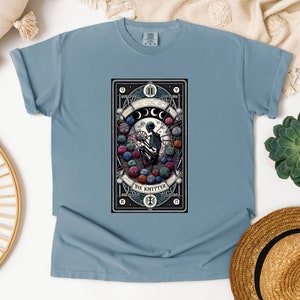 May include: A light blue t-shirt with a graphic of a tarot card depicting a skeleton knitting with yarn balls. The card is titled "The Knitter" and has Roman numerals "XI" at the top.
