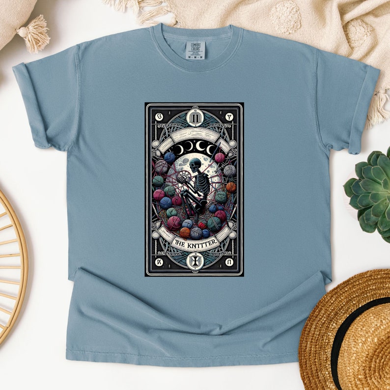 May include: A light blue t-shirt with a graphic of a tarot card depicting a skeleton knitting with yarn balls. The card is titled "The Knitter" and has Roman numerals "XI" at the top.