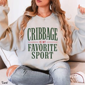 May include: A sand-colored crewneck sweatshirt featuring the dark green text "CRIBBAGE" above the red text "IS MY" and the dark green text "FAVORITE SPORT" centered on the chest. The garment has long sleeves and a ribbed collar and cuffs.