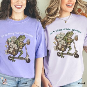 May include: Two light purple t-shirts with the text "OFF TO LEGALLY DOCUMENT EVERYTHING". Each shirt features a cartoon frog riding a scooter, carrying a briefcase, and holding a coffee. The shirt on the left is violet, and the shirt on the right is orchid.