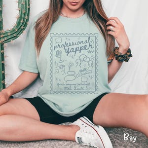 以下が含まれることがあります： 水色のTシャツ。青色のグラフィックは、切手のような枠の中に「professional yapper」と書かれています。グラフィックには、マイク、吹き出し、その他のスピーチ関連のシンボルが含まれています。「speech language pathologist Mrs. Smith」というテキストも含まれています。