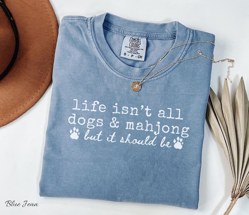 May include: A blue jean colored short-sleeve t-shirt with a crew neckline. The shirt features white printed text that reads, life isn't all dogs & mahjong but it should be, accompanied by two small white paw print graphics below the text.
