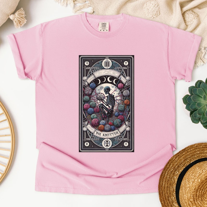 May include: Pink t-shirt with a graphic of a tarot card featuring a skeleton knitting with yarn balls. The card is titled "The Knitter".