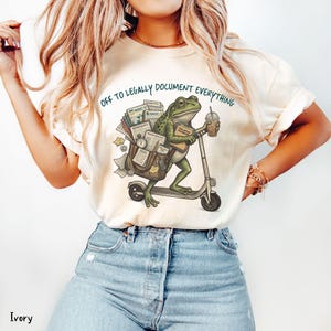 May include: Ivory-colored t-shirt with a whimsical illustration of a frog riding a scooter, carrying documents and a coffee cup. The text "OFF TO LEGALLY DOCUMENT EVERYTHING" is printed above the frog. The shirt is paired with blue jeans.