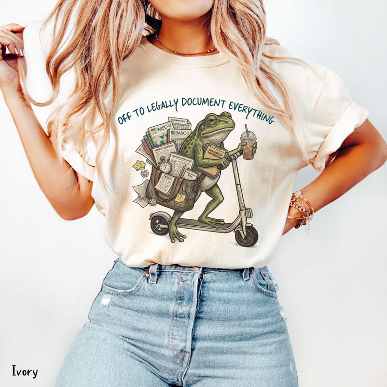 May include: Ivory-colored t-shirt with a whimsical illustration of a frog riding a scooter, carrying documents and a coffee cup. The text "OFF TO LEGALLY DOCUMENT EVERYTHING" is printed above the frog. The shirt is paired with blue jeans.