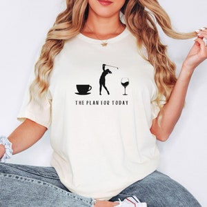Op de afbeelding: Een wit T-shirt met een zwarte afbeelding van een vrouw die golf speelt, een kopje koffie en een glas wijn. De tekst "THE PLAN FOR TODAY" staat onder de afbeelding.