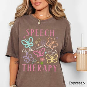 Puede incluir: Camiseta marr&oacute;n con las palabras "SPEECH THERAPY" en rosa y la frase "helps little voices take flight". El dise&ntilde;o incluye gr&aacute;ficos de mariposas de colores en verde azulado, rosa, naranja, morado y amarillo.