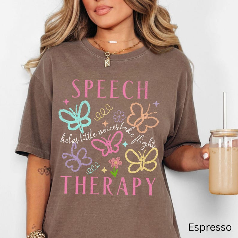 Puede incluir: Camiseta marr&oacute;n con las palabras "SPEECH THERAPY" en rosa y la frase "helps little voices take flight". El dise&ntilde;o incluye gr&aacute;ficos de mariposas de colores en verde azulado, rosa, naranja, morado y amarillo.