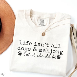 May include: An ivory short-sleeve t-shirt with a crew neckline and a Comfort Colors brand label. The shirt features black printed text that reads, "life isn't all dogs & mahjong but it should be" accompanied by two small black paw print graphics.
