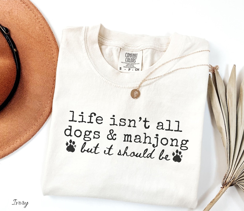 May include: An ivory short-sleeve t-shirt with a crew neckline and a Comfort Colors brand label. The shirt features black printed text that reads, "life isn't all dogs & mahjong but it should be" accompanied by two small black paw print graphics.