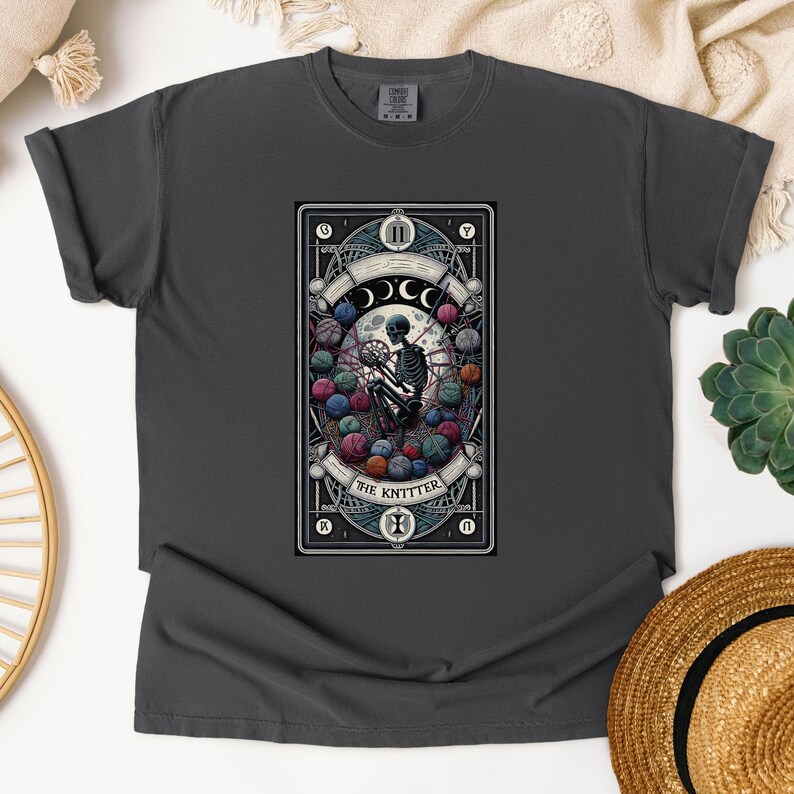 May include: A dark gray t-shirt with a graphic of a tarot card featuring a skeleton knitting with yarn balls. The card is titled "The Knitter".