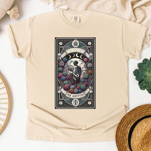 May include: A beige t-shirt with a graphic of a tarot card featuring a skeleton knitting with yarn balls. The card is titled "The Knitter" and has a black background with gold accents.