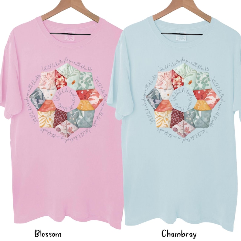 May include: Two t-shirts with a colorful patchwork design in the center. The design is a circle with the text "I still like to play with blocks" written around the outside. The t-shirt on the left is pink and the t-shirt on the right is light blue.