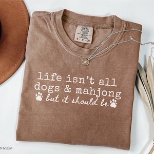 May include: A brown short-sleeved t-shirt with a crew neckline featuring the white printed text 'life isn't all dogs & mahjong but it should be' accompanied by two paw print icons. The shirt has a Comfort Colors brand label inside the collar.