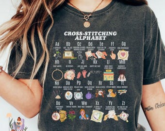 May include: A black t-shirt with a white graphic print of a cross-stitching alphabet. The graphic includes a letter and a cross-stitch design for each letter of the alphabet. The text "CROSS-STITCHING ALPHABET" is at the top of the graphic.