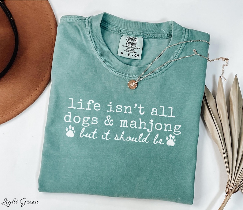 May include: A light green short-sleeve t-shirt with a crew neckline and a white graphic print that reads, "life isn't all dogs & mahjong but it should be" accompanied by two paw print icons. The shirt features a Comfort Colors brand neck label.