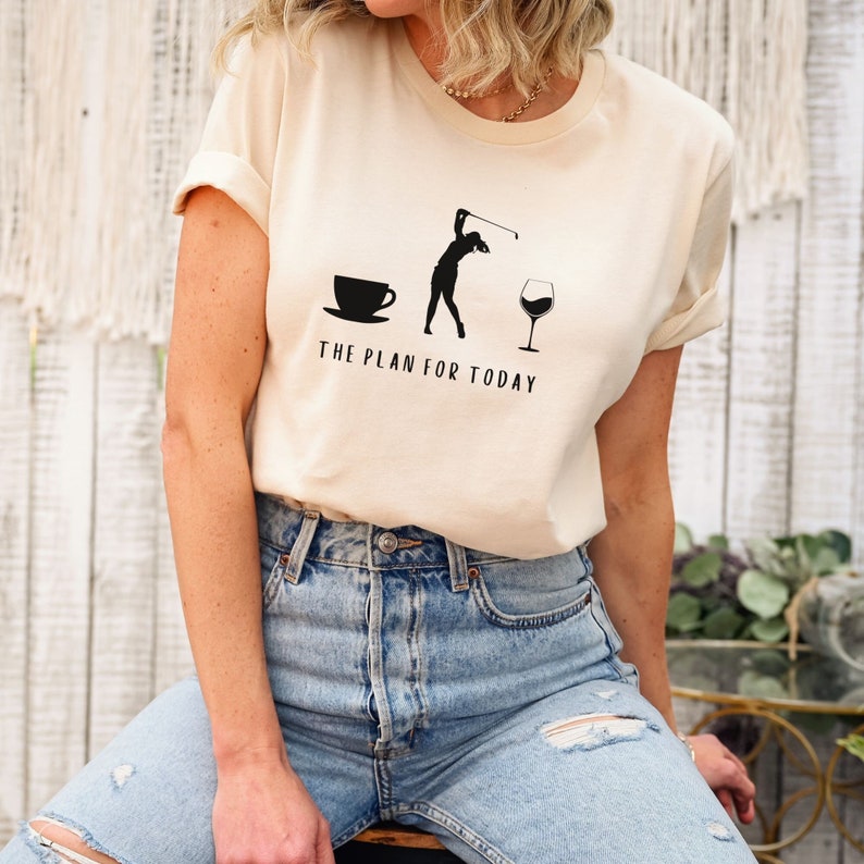 Op de afbeelding: Een beige T-shirt met een afbeelding van een golfer, een kopje koffie en een glas wijn. De tekst "THE PLAN FOR TODAY" staat onder de afbeelding.