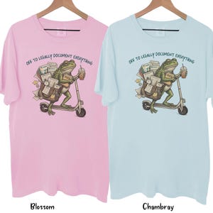 May include: Two t-shirts, one pink and one light blue, each with a graphic of a frog riding a scooter. The frog carries documents and a coffee cup. Text on the shirts reads "OFF TO LEGALLY DOCUMENT EVERYTHING."