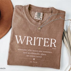 May include: A brown short-sleeved t-shirt with white text that reads WRITER noun, someone who hears one sentence and accidentally plans an entire novel, est. 3 a.m. The shirt features a crew neckline and a Comfort Colors brand label inside.