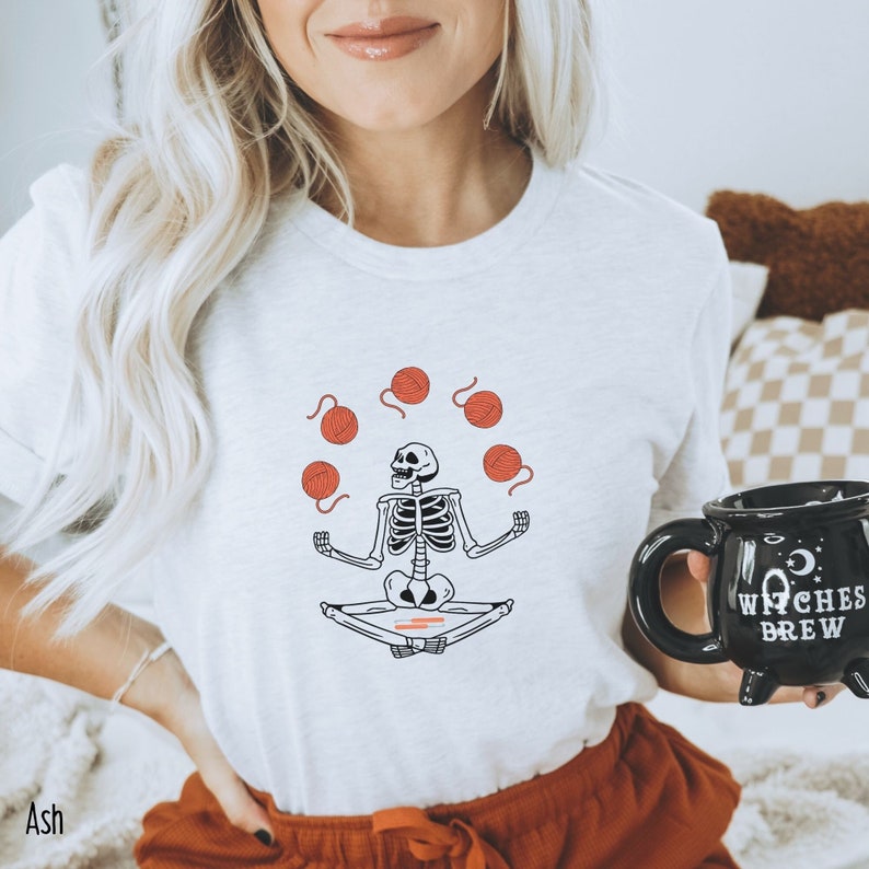 May include: A white t-shirt with a graphic of a skeleton meditating and juggling balls of yarn. The text "Witches Brew" is on a black mug in the background.