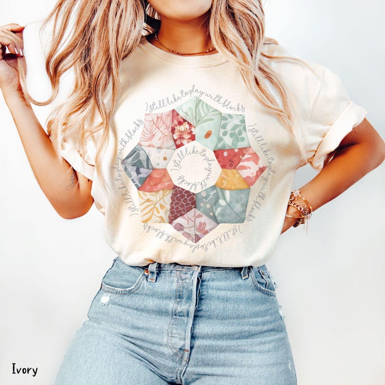 May include: A white t-shirt with a circular design of colorful fabric squares. The design is surrounded by the text "I still like to play with blocks".