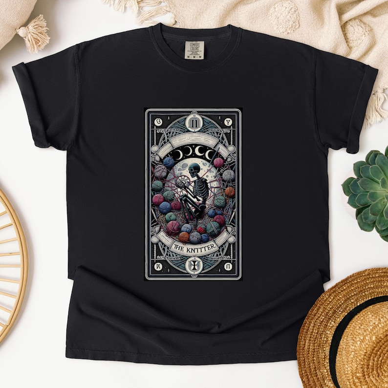 May include: Black t-shirt with a graphic of a tarot card featuring a skeleton knitting with yarn balls. The card is titled "The Knitter".
