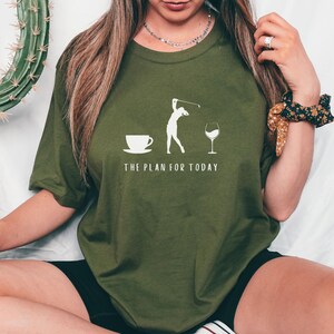 Op de afbeelding: Een groen T-shirt met een witte afbeelding van een vrouw die golf speelt, een kopje koffie en een glas wijn. De tekst "THE PLAN FOR TODAY" staat onder de afbeelding.