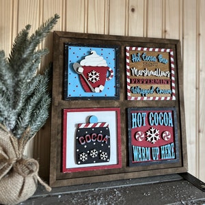 May include: A wooden sign with four squares, each with a different design. The top left square has a red and white mug with whipped cream and a snowflake on a blue background with white polka dots. The top right square has a red and white striped border with the text "Hot Cocoa Bar" in red and white letters. The text "MENU" is in white letters. The text "Marshmallows!" is in red letters. The text "PEPPERMINT" is in red letters. The text "Whipped Cream" is in red letters. The bottom left square has a red and white striped jar with the text "COCOA" in white letters. The bottom right square has a red and white striped border with the text "HOT COCOA" in red and white letters. A snowflake is in the center of the square. The text "WARM UP HERE" is in red and white letters.
