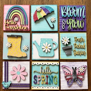 May include: Nine wooden signs with a spring theme. The signs feature a rainbow, an umbrella, a boot, a watering can, a flower pot, a flower, a butterfly, a bird, and the words "Hello Sunshine", "Bloom & Grow", "Fresh Picked Daisies", "hello SPRING".