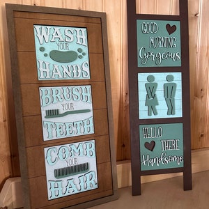 May include: Two wooden signs with a bathroom theme. The first sign has a light blue background with the words "Wash Your Hands", "Brush Your Teeth", "Comb Your Hair" in white. The second sign has a light blue background with the words "Good Morning Gorgeous", "Hello There Handsome" in white.