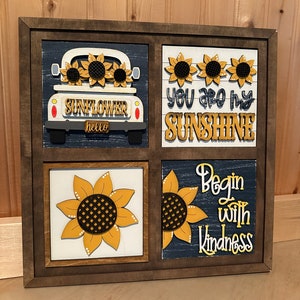 May include: A wooden framed wall art piece with four squares. Each square features a sunflower design and a different saying. The top left square shows a white pickup truck with sunflowers in the bed and the words "SUNFLOWER hello". The top right square has three sunflowers and the words "You are my SUNSHINE". The bottom left square has a single sunflower. The bottom right square has a sunflower and the words "Begin with Kindness".