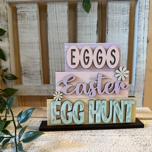 May include: A three-tiered wooden sign with pastel colors. The top tier is lavender and says "EGGS". The middle tier is light pink and says "Easter". The bottom tier is light blue and says "EGG HUNT".