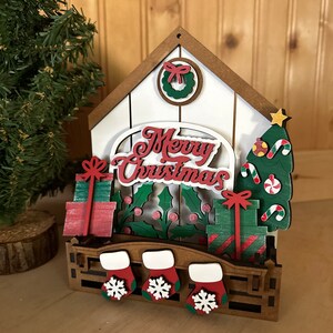 May include: Wooden Christmas house decoration with a white background, red and green accents, and the words "Merry Christmas". The house has a wreath, presents, a Christmas tree, and stockings.