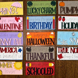 May include: A collection of colorful wooden signs with various seasonal and holiday-themed phrases. Phrases include "Welcome Spring," "Happy Halloween," and "True North." Each sign is rectangular and features a unique design and color scheme.