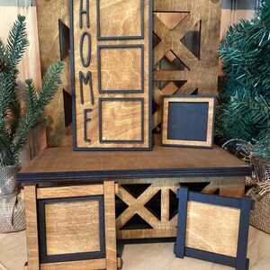 May include: Wooden sign with the word "HOME" spelled vertically in black lettering. The sign has three rectangular spaces for photos or other decorations. The sign is on a wooden shelf with a black frame. There are two additional square wooden frames with black borders on the shelf.