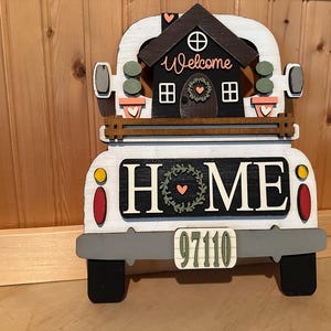 May include: White wooden truck with a black house on the back. The house has a welcome sign and a wreath on the door. The truck has a black and white sign that says "HOME" and a license plate that says "97110".