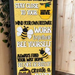 May include: A yellow wooden sign with black lettering and bee-themed decorations. The sign reads "Advice from a honey bee, Stay close to your hive, Mind your own beeswax, Work together, Bee yourself, Always find your way home, Bee nice or buzz off, Create a buzz".