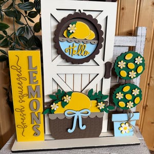 May include: A layered wooden decorative sign featuring a white door with a brown circular wreath displaying a yellow lemon and the word Hello. To the left is a yellow sign reading fresh squeezed LEMONS. To the right is a potted lemon tree.