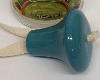 May include: A teal ceramic bottle stopper with a white wick. The stopper has a smooth, glossy finish, with a rounded top and flared base. The wick is made of natural fibre. A jar of food is in the background.