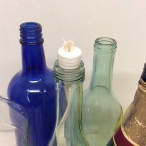 Op de afbeelding: Vier glazen flessen, waarvan één met een witte keramische bovenkant en een lont, klaar om te worden gebruikt als een decoratieve kaarsenhouder.