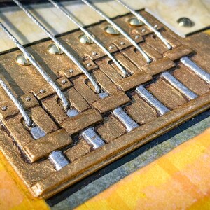 Puede incluir: Un puente de guitarra con una base de metal texturizado de color dorado y seis selletas plateadas individuales. El puente est&aacute; montado sobre un cuerpo de guitarra a rayas amarillas claras y naranjas, con seis cuerdas plateadas sobre las selletas.