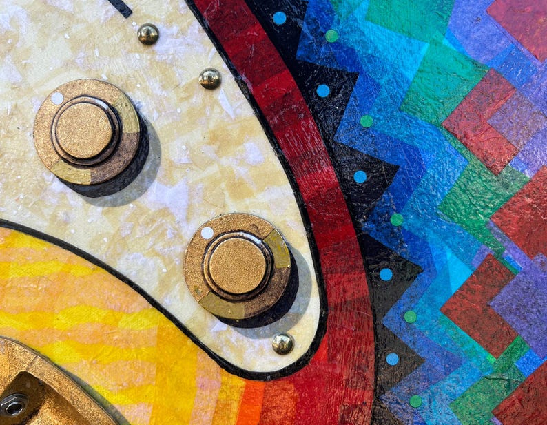 Puede incluir: Un primer plano de una superficie texturizada con dos perillas circulares doradas montadas sobre un panel color crema con brillo met&aacute;lico. A la derecha, un patr&oacute;n geom&eacute;trico vibrante muestra l&iacute;neas en zigzag rojas, negras, azules y verdes.