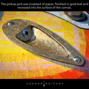 Puede incluir: Un jack de entrada esculpido en papel con acabado de pan de oro, empotrado en una superficie de lienzo con patrones amarillos y naranjas. El texto dice: The pickup jack was sculpted of paper, finished in gold leaf and recessed into the surface of the canvas.