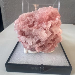 May include: A cluster of translucent, pale pink cubic mineral crystals resting on a white base inside a clear plastic display box with a black frame. The crystalline structure features sharp, geometric edges and a textured, multifaceted surface.