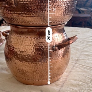 Pode incluir: Uma panela de cozinha cor de cobre com tampa, com uma textura martelada. A panela tem aproximadamente 29 cm de altura e duas alças. A tampa tem uma pequena alça na parte superior. A panela está sobre uma toalha de mesa branca.