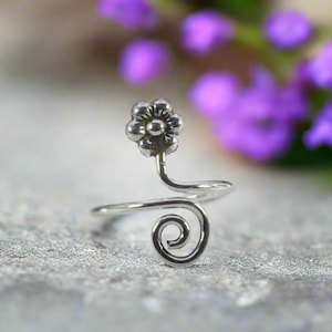 May include: A silver ring with a flower motif. The ring has a small flower at the top and a spiral design at the base. The background is blurred, showing purple flowers.