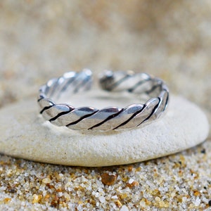 Pode incluir: Um anel de dedo do pé de prata com um design trançado. O anel está sobre uma rocha branca.