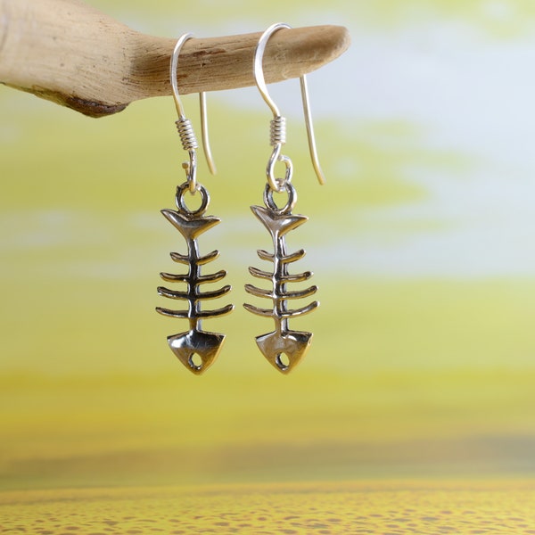 May include: Silver fish bone earrings hanging from a piece of driftwood. The earrings are silver and have a detailed fish bone design.