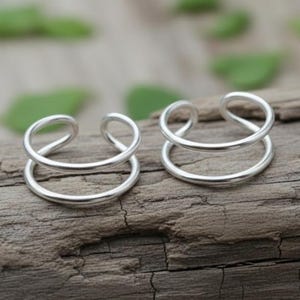 May include: Two silver-colored double-band ear cuffs. Each cuff has two circular bands connected by a curved top. The jewelry is displayed on a weathered, gray wooden surface, with green foliage in the background.